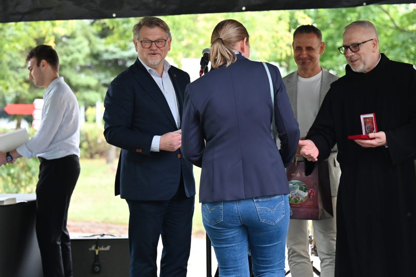 Zakonnik w czarnym habicie wręcza medal kobiecie w granatowej marynarce. Obok stoją mężczyźni w garniturach i uśmiechają się podczas ceremonii