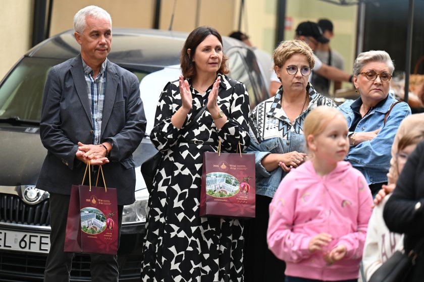 Grupa osób stojących przed samochodem, obserwujących wydarzenie. Mężczyzna w szarej marynarce i kobieta w czarno-białej sukience trzymają torby jubileuszowe Bonifratrów