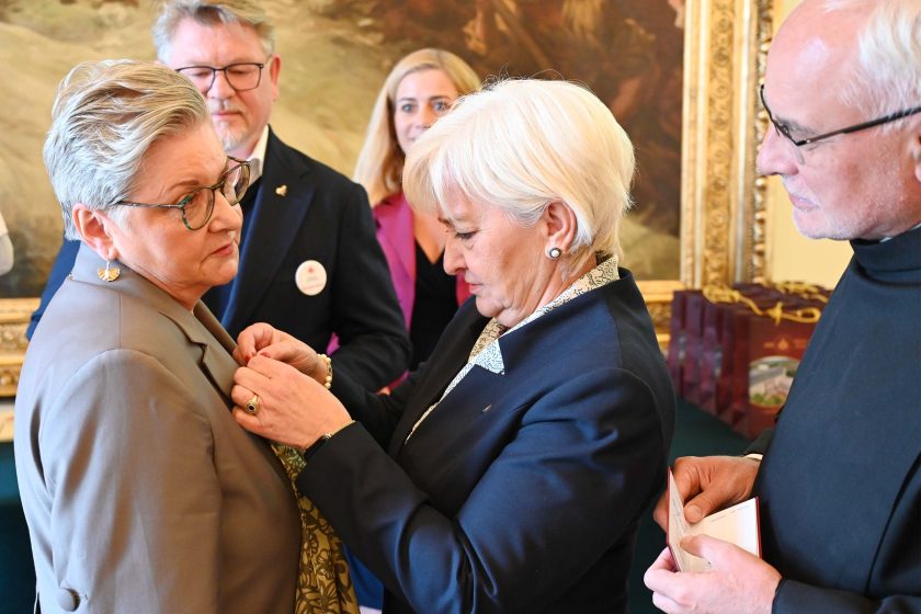 Zbliżenie sceny dekoracji: kobieta o krótkich, białych włosach w granatowym kostiumie przypina metalowe odznaczenie na lewej klapie szarej marynarki starszej kobiety w okularach. Odznaczana ma złote kolczyki i wzorzystą, oliwkowo-złotą apaszkę przewieszoną przez szyję. W tle widać mężczyznę w granatowej marynarce z przypinką-okrągłą plakietką, kobietę w malinowej marynarce oraz zakon­nika w czarnym habicie trzymającego otwarte etui lub dokument. Tło stanowi duży obraz w bogato rzeźbionej, złotej ramie i rząd bordowych toreb upominkowych na zielonym obrusie.