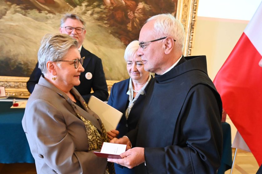 Po akcie dekoracji odznaczona kobieta w szarym żakiecie stoi naprzeciw zakonnika w czarnym habicie; wymieniają kilka słów i dokumenty w bordowych okładkach. Za nimi uśmiechnięta kobieta dekorująca (białe włosy, granatowy kostium) oraz mężczyzna w granatowej marynarce. Po prawej fragment stojącej polskiej flagi. Tłem jest monumentalny obraz w złotej ramie.