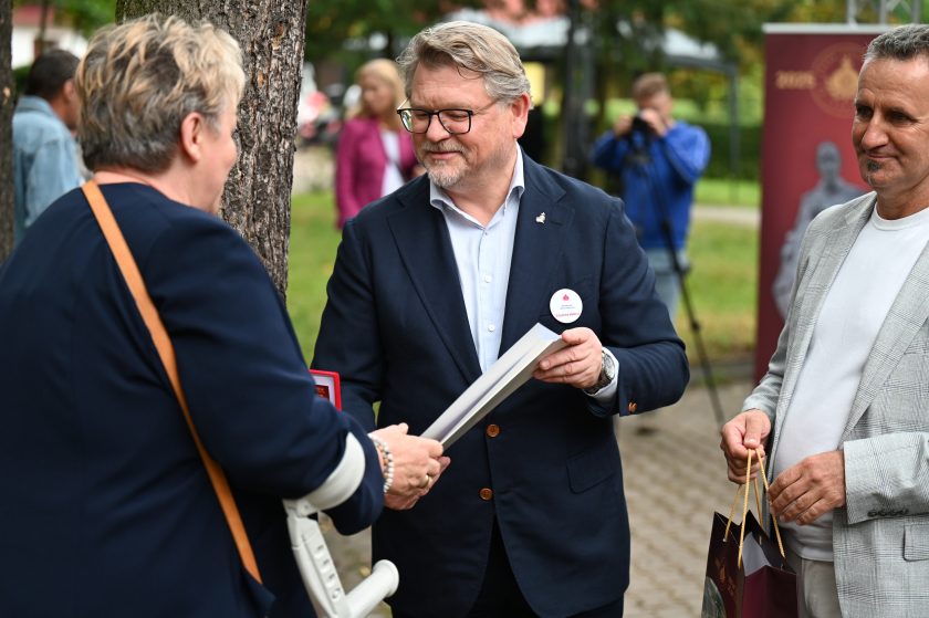 Mężczyzna w granatowej marynarce wręcza nagrodę kobiecie opartej na kuli ortopedycznej. Uśmiechnięci uczestnicy stoją obok, trzymając torby jubileuszowe
