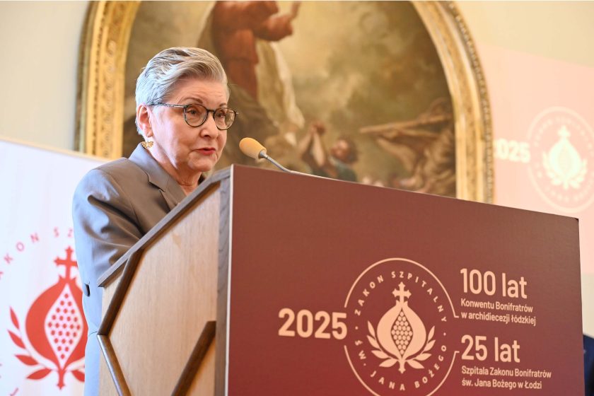 Odznaczona kobieta wygłasza krótkie przemówienie zza mównicy z bordowym frontem, na którym widnieje duże koło z emblematem w kształcie granatu i napisy jubileuszu: „2025”, „100 lat Konwentu Bonifratrów w archidiecezji łódzkiej”, „25 lat Szpitala Zakonu Bonifratrów św. Jana Bożego w Łodzi”. Kobieta ma szary żakiet i okrągłe okulary; mówi do mikrofonu na giętym, jasnym wysięgniku. W tle rozmyty owalny obraz religijny w złotej ramie oraz fragment roll-upu z czerwonym symbolem zakonu.