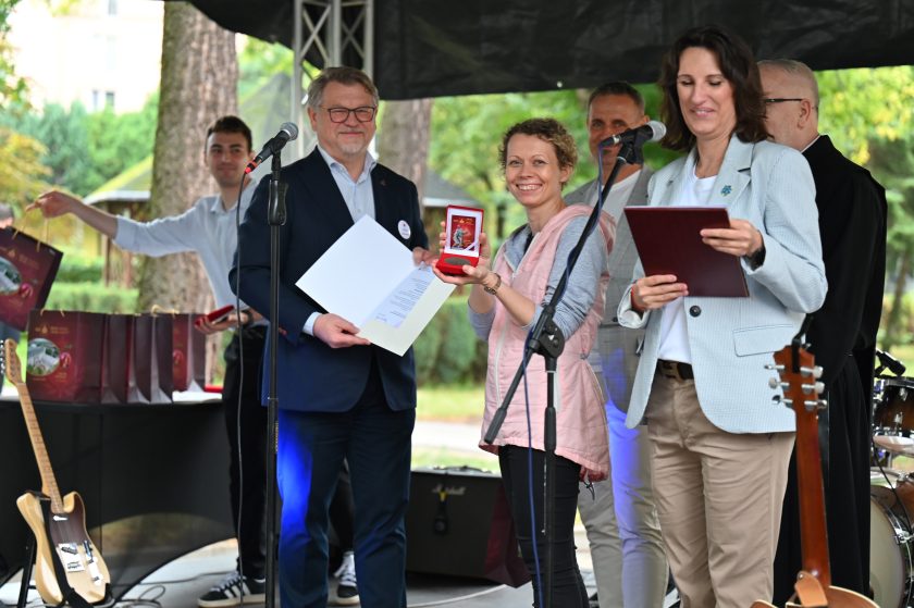 Kobieta w różowej kamizelce prezentuje medal w czerwonym etui, uśmiechając się szeroko do obiektywu. Obok niej prowadząca uroczystość czyta dokument