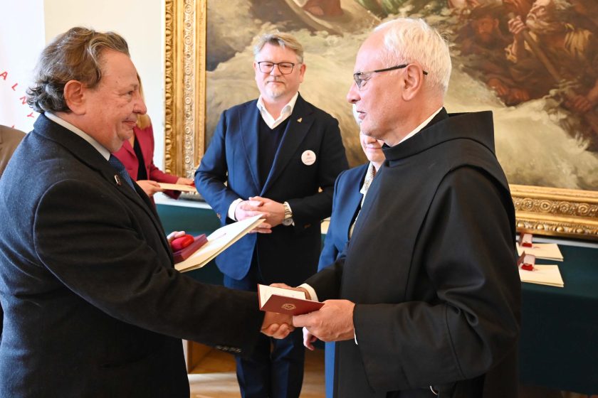 Ujęcie z wręczenia: starszy mężczyzna w ciemnym płaszczu i niebieskim krawacie ściska dłoń zakonnika w czarnym habicie. Obaj trzymają dokumenty oraz bordowe etui na medal. Za nimi stoją organizatorzy — mężczyzna w granatowej marynarce i kobieta w granatowym kostiumie — na stole w tle leżą białe teczki i pudełka z odznaczeniami. Ścianę wypełnia wielki obraz z dynamiczną sceną.