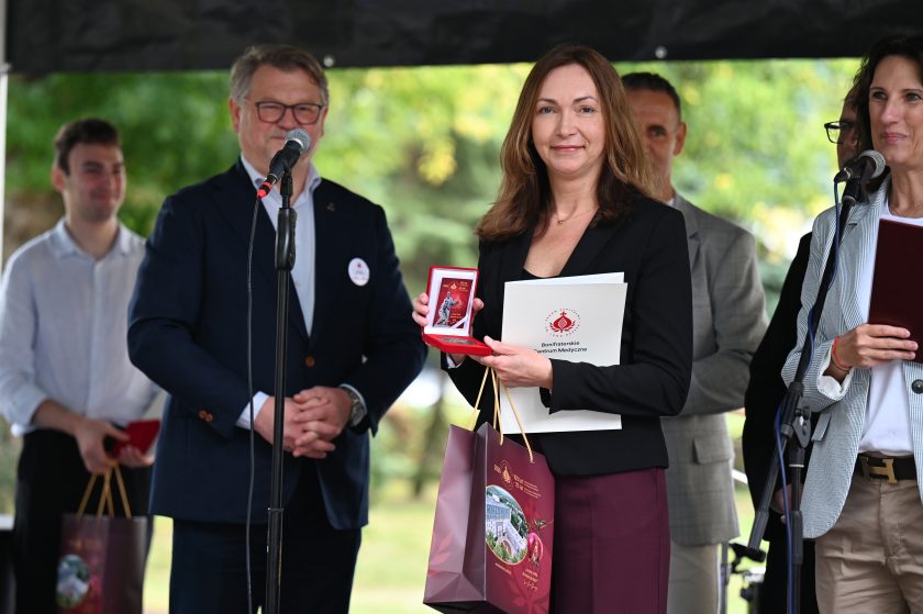 Kobieta w ciemnym żakiecie trzyma medal i dyplom Bonifraterskiego Centrum Medycznego. Za nią mężczyzna w garniturze uśmiecha się w stronę aparatu