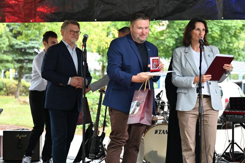 Mężczyzna w granatowej marynarce otrzymuje medal i dyplom na scenie. W ręku trzyma też torbę jubileuszową Bonifratrów