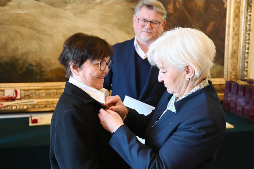 Kobieta o białych włosach przypina medal na klapie czarnego żakietu starszej kobiety w okularach i białej koszuli. Odznaczana uśmiecha się lekko, spoglądając w dół. W tle mężczyzna w granatowej marynarce obserwuje scenę; po prawej rząd bordowych toreb upominkowych na zielonym obrusie. Po lewej kadr obejmuje dłoń z czerwonym lakierem na paznokciach trzymającą następne etui i dyplom.