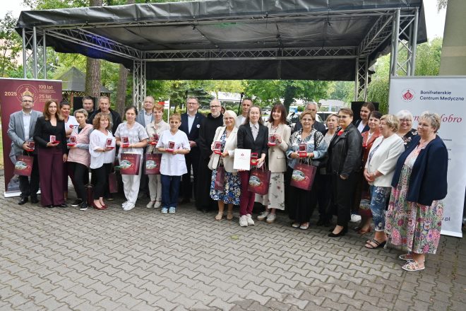 Zdjęcie grupowe laureatów i uczestników wydarzenia. Kilkanaście osób stoi przed sceną z medalami i torbami jubileuszowymi w rękach