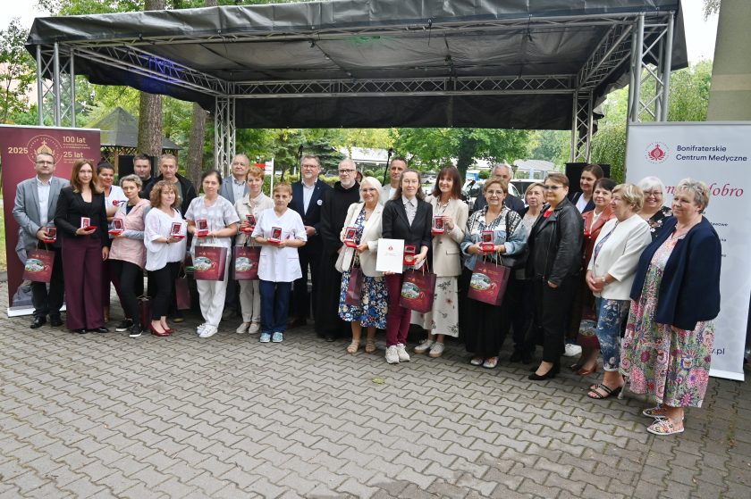 Zdjęcie grupowe laureatów i uczestników wydarzenia. Kilkanaście osób stoi przed sceną z medalami i torbami jubileuszowymi w rękach