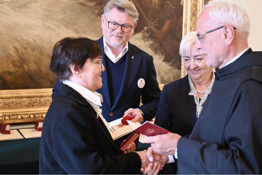 Moment gratulacji po dekoracji: odznaczona kobieta w czarnym żakiecie ściska dłoń zakonnika w habicie. Z boku widać mężczyznę w granatowej marynarce z dyplomem oraz kobietę dekorującą, która uśmiecha się i obserwuje uścisk. W dłoniach i na stole widoczne bordowe pudełka na medale; za nimi złota rama obrazu wypełniającego tło.