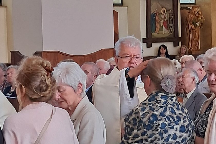 Kapłan w białej szacie udziela błogosławieństwa kobiecie podczas nabożeństwa, w tle widać wiernych modlących się w ławkach