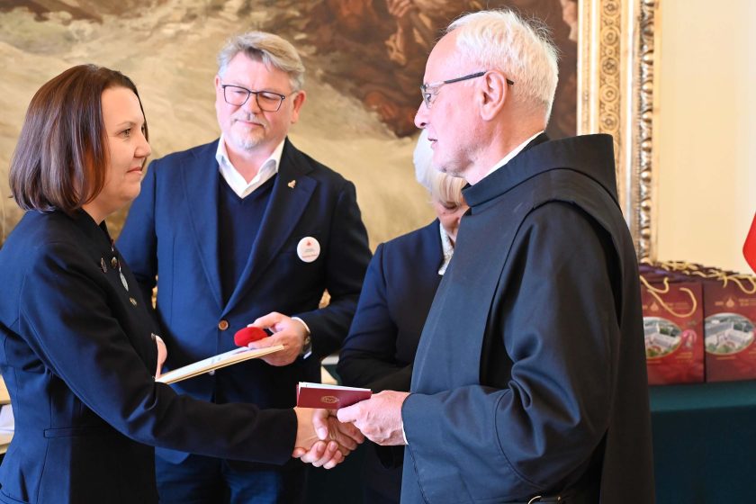 Wręczenie dokumentów kolejnej odznaczonej: kobieta o brązowych, do ramion włosach, w ciemnej, dwurzędowej marynarce, podaje dłoń zakonnikowi w czarnym habicie. Mężczyzna w granatowej marynarce trzyma obok bordowe etui i dyplom; kobieta dekorująca stoi częściowo zasłonięta. W tle rząd bordowych toreb prezentowych i fragment polskiej flagi.
