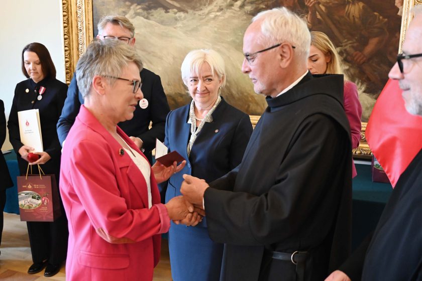 Kobieta w różowym żakiecie przyjmuje gratulacje — uściskiem dłoni i wręczeniem etui na medal — od mężczyzny w czarnym habicie. Obok stoi uśmiechnięta organizatorka w granatowym kostiumie; w tle wielkoformatowy obraz i osoby oczekujące z teczkami oraz torbami upominkowymi.