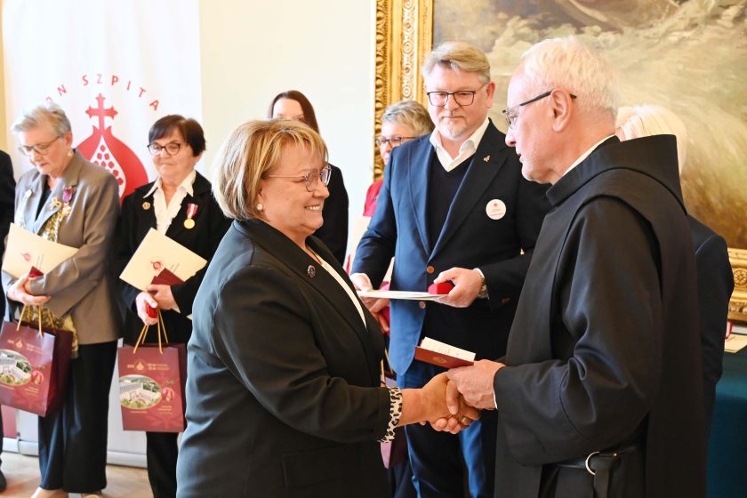 Uśmiechnięta kobieta w ciemnym kostiumie ściska dłoń zakonnika w czarnym habicie podczas wręczania odznaczenia. Za nią sznur osób z dyplomami; widoczny mężczyzna w granatowej marynarce trzyma dokument i czerwone etui. Sceneria sali: jasne ściany, złocona rama obrazu.