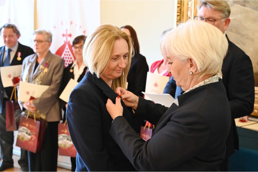 Kobieta z krótkimi blond włosami w granatowej marynarce otrzymuje przypinkę — druga kobieta, o białych włosach, przypina medal do klapy jej żakietu. W tle rząd odznaczonych z teczkami, roll-up z logo i bordowe torby ustawione na stole.