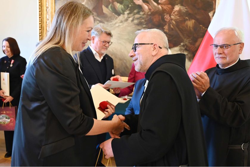 Wysoka kobieta w czarnym żakiecie i białej bluzce ściska dłoń uśmiechniętego zakonnika w czarnym habicie i okularach; w drugiej ręce trzyma czerwone etui z odznaczeniem i kopertę. Za nimi drugi zakonnik bije brawo. Tło stanowią obraz w złotej ramie i polska flaga.