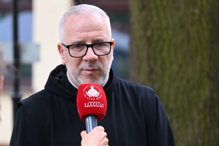 Zakonnik w czarnym habicie udziela wywiadu na tle drzewa. Przed nim trzymany mikrofon z czerwonym logo Fundacji Bonifraterskiej