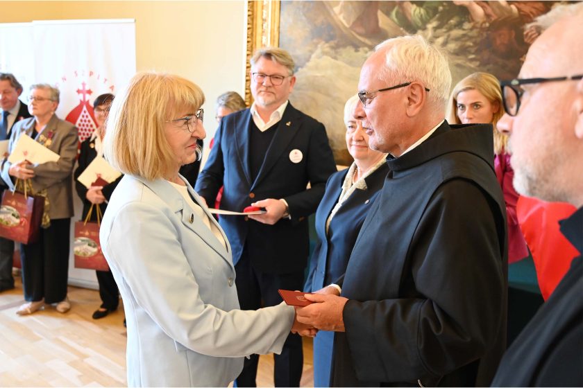 Kobieta w jasnobłękitnym żakiecie i okularach ściska dłoń zakonnika w czarnym habicie, który przekazuje czerwone etui i dokument. W tle organizatorzy: mężczyzna w granatowej marynarce i kobieta w granatowym kostiumie; za nimi roll-up z logo zakonu.