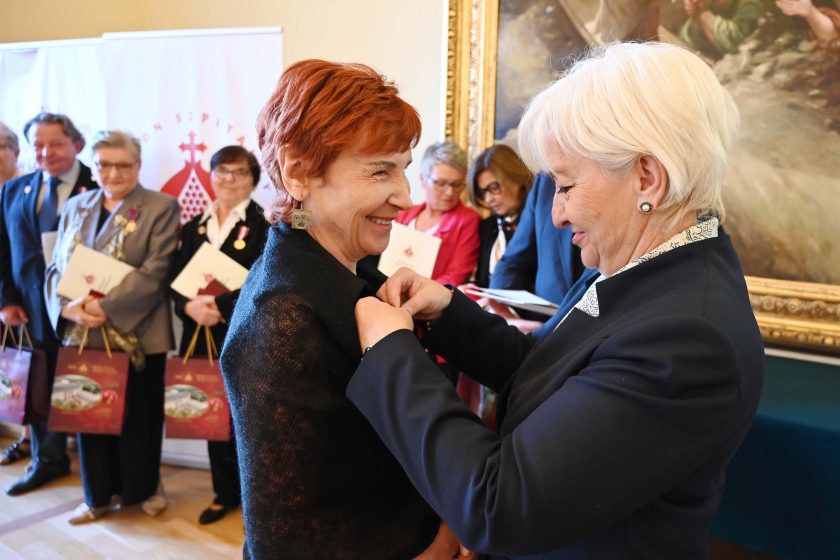 Kobieta o krótkich rudych włosach, w czarnym swetrze, uśmiecha się, gdy organizatorka o białych włosach przypina jej medal do lewej klapy. W tle kolejka odznaczonych z dyplomami i bordowymi torbami prezentowymi; kadr bliski, emocje radości i skupienia.