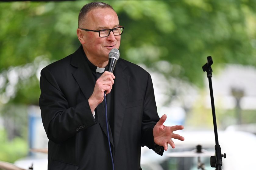Ksiądz w czarnej marynarce i koloratce przemawia do mikrofonu na scenie plenerowej. Gestykuluje dłonią, a w tle rozmyta zieleń drzew