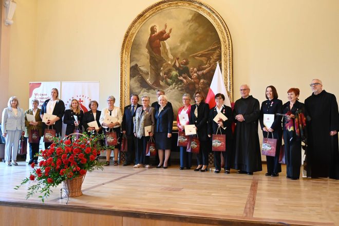 Kilkanaście osób stoi w jednym rzędzie na scenie sali uroczystości; w tle ogromny obraz religijny w złotej ramie oraz dwa roll-upy z napisem „Wyższe Seminarium Duchowne w Łodzi” i logo Zakonu. Większość trzyma dyplomy w kremowych teczkach i bordowe torby z upominkami. Po prawej stoi biało-czerwona flaga na maszcie, a na proscenium kosz z czerwonymi goździkami.