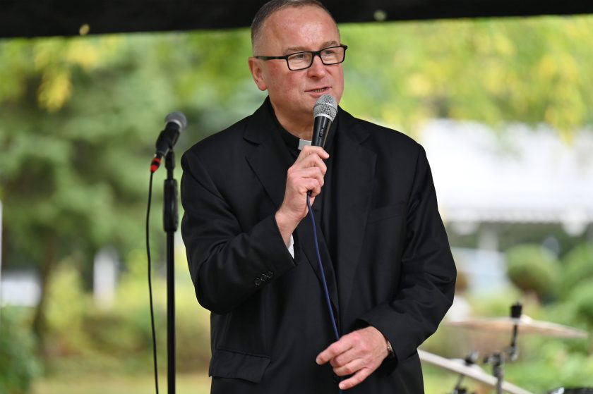 Ksiądz w czarnej marynarce i koloratce trzyma mikrofon i mówi podczas wystąpienia. W tle widoczna perkusja ustawiona na scenie