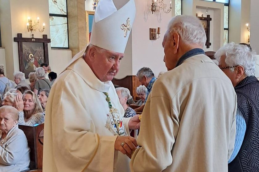 Biskup w białych szatach liturgicznych podaje komunię starszemu mężczyźnie, w tle pozostali wierni uczestniczą w Eucharystii