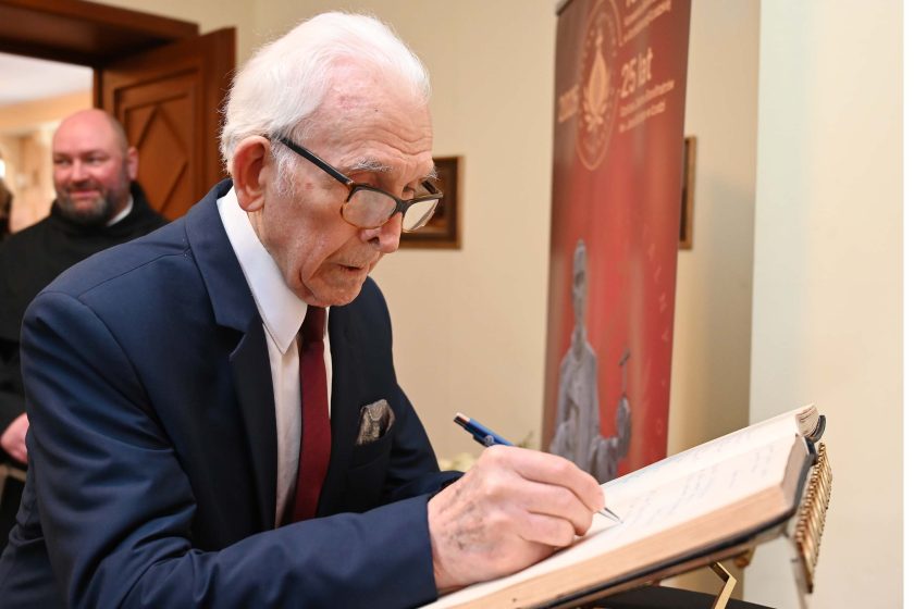 Starszy mężczyzna w garniturze i czerwonym krawacie wpisuje się do księgi pamiątkowej jubileuszu.