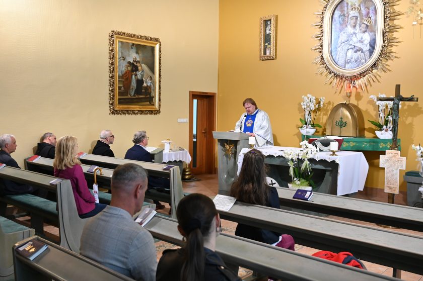 Widok ogólny wnętrza kaplicy. Kapłan w srebrnym ornacie głosi kazanie z ambony, a wierni siedzą w ławkach i słuchają. Na ścianie obraz Matki Bożej z Dzieciątkiem oraz inny obraz przedstawiający scenę biblijną