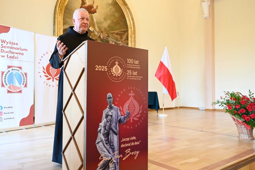 Mężczyzna w czarnym habicie przemawia przy mównicy. Front mównicy jest bordowy z dużym logo „2025 / 100 lat / 25 lat” i grafiką rzeźby św. Jana Bożego oraz cytatem. Po prawej pusta przestrzeń sceny, flaga Polski i kosz czerwonych kwiatów; po lewej roll-upy organizatora.