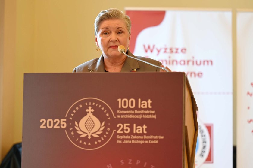 Kobieta o siwych, krótko ściętych włosach przemawia zza dużej mównicy z bordowym frontem. Na pulpicie widoczny znak „Zakon Szpitalny św. Jana Bożego” oraz napisy: „2025”, „100 lat Konwentu Bonifratrów w archidiecezji łódzkiej”, „25 lat Szpitala Zakonu Bonifratrów św. Jana Bożego w Łodzi”. W tle roll-upy seminarium duchownego. Mówczyni ma szarą marynarkę i złote kolczyki, mówi do mikrofonu.