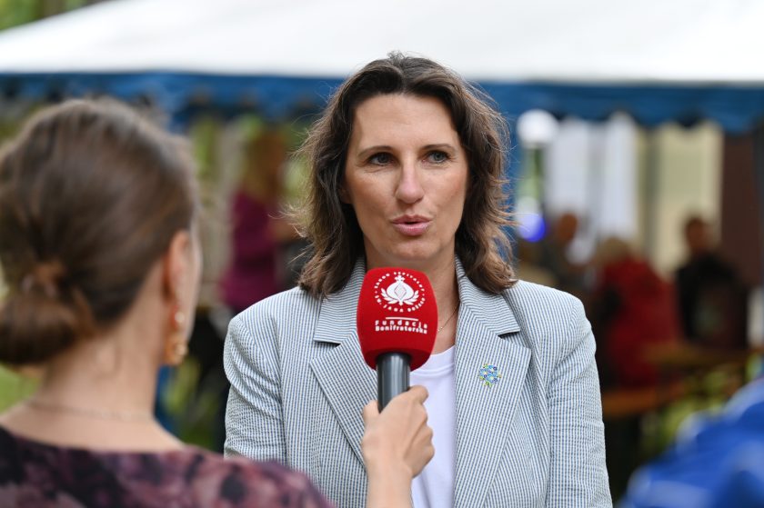 Kobieta w jasnej marynarce udziela wywiadu. Przed nią mikrofon z czerwonym logo Fundacji Bonifraterskiej, w tle widoczni uczestnicy spotkania