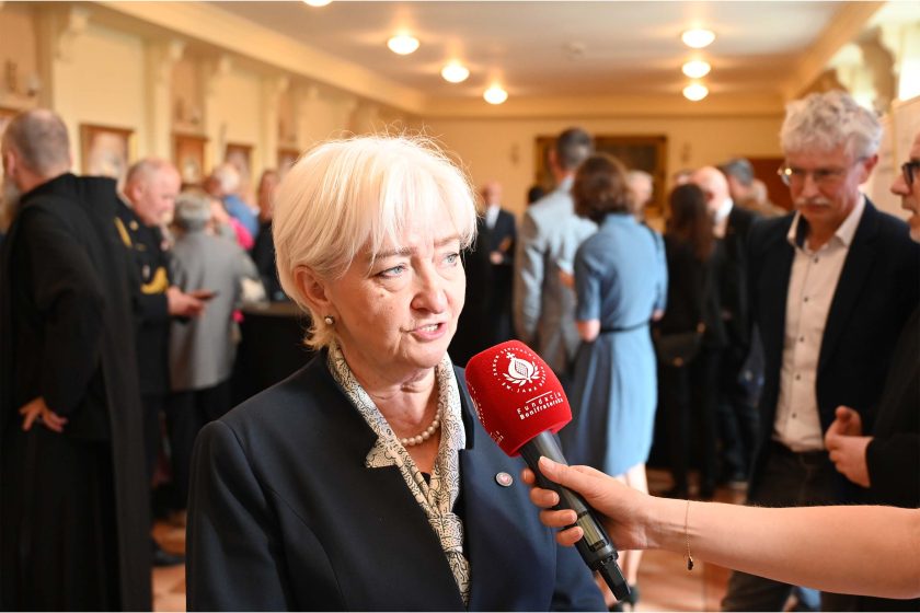 Wywiad w korytarzu – kobieta o białych włosach w granatowym kostiumie i perłowym naszyjniku odpowiada do czerwonego mikrofonu z logo „Fundacja Bonifratri”. Za nią tłum gości rozmawiających w półcieniu, ciepłe światło sufitowych plafonów.