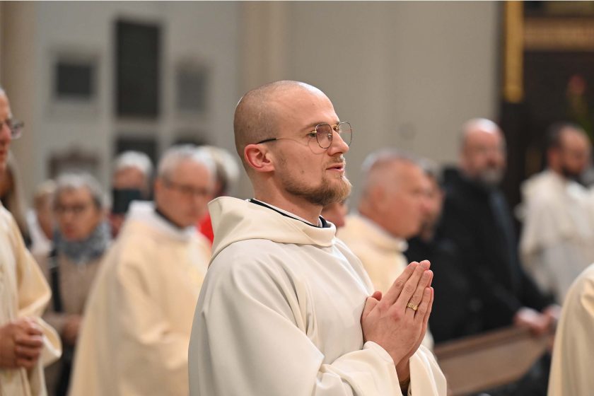 Młody zakonnik z ogoloną głową i krótką bródką, w okularach, stoi w ławce złożony do modlitwy – dłonie z obrączką na palcu złączone przy piersi. Ubrany w jasny habit z kapturem; w tle rozmyci kapłani i wierni.
