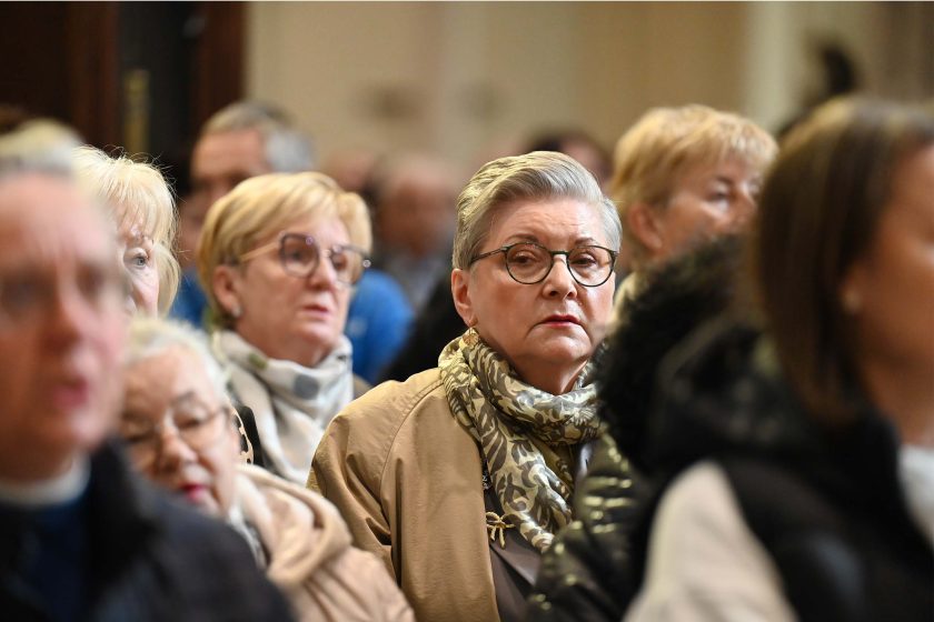 Zbliżenie na kobietę w okrągłych okularach pośród wiernych; ma beżowy płaszcz i wzorzysty, oliwkowo-beżowy szal. Jej twarz jest ostra i dobrze oświetlona, tło innych osób pozostaje miękko rozmyte.
