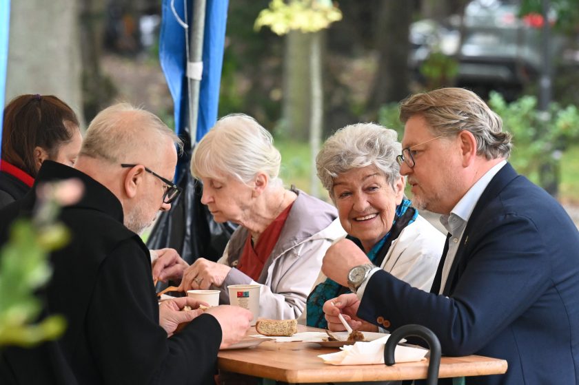 Grupa osób siedzi przy stole plenerowym, jedząc posiłek. Starsza kobieta uśmiecha się szeroko, rozmawiając z zakonnikiem i mężczyzną w garniturze
