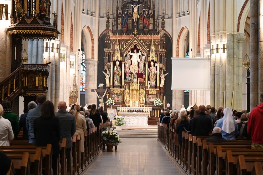 Szeroki kadr wnętrza kościoła z długą nawą, ławkami po obu stronach i wiernymi stojącymi podczas liturgii. Na osi bogato złocony, piętrowy ołtarz z figurą Zmartwychwstałego w centrum; po lewej ambona, po prawej zwinięty ekran projekcyjny. Przy prezbiterium kwiaty i obrazy, a witraże rzucają kolorowe refleksy.
