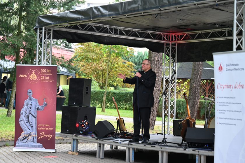 Ksiądz w czarnej marynarce przemawia na scenie plenerowej. Obok stoją banery jubileuszowe Zakonu Bonifratrów