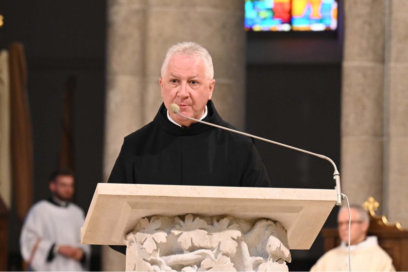 Kapłan w czarnym habicie głosi homilię przy kamiennym ambonie z rzeźbionymi liśćmi; przed nim cienki mikrofon na długim wysięgniku. Twarz oświetlona ciepłym światłem, za nim rozmyte tło nawy i fragment witraża. Z boku niewyraźna sylwetka innego celebransa siedzącego w stallach.