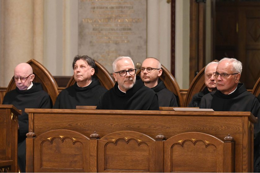 Rząd duchownych w czarnych habitach siedzących w rzeźbionych, drewnianych stallach. Sześciu mężczyzn, część w okularach, o poważnych, skupionych minach; środkowy lekko zwrócony w lewo. Za nimi jasny kamień i tablice pamiątkowe. Oświetlenie równomierne, podkreślające ciepły kolor drewna.