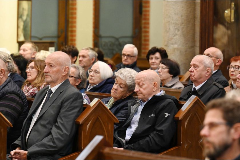 Ujęcie boczne na grupę wiernych; wśród nich mężczyzna z kręconymi, jasnymi włosami i okularami, obok starsza kobieta o białych włosach. Wszyscy siedzą, patrząc w jedną stronę, twarze skupione. W tle masywne kolumny i ciemne płyty ścienne. Kadr równoważy chłód kamienia z ciepłem światła wnętrza.
