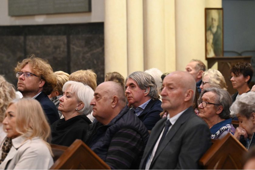 Ujęcie boczne na grupę wiernych; wśród nich mężczyzna z kręconymi, jasnymi włosami i okularami, obok starsza kobieta o białych włosach. Wszyscy siedzą, patrząc w jedną stronę, twarze skupione. W tle masywne kolumny i ciemne płyty ścienne. Kadr równoważy chłód kamienia z ciepłem światła wnętrza.