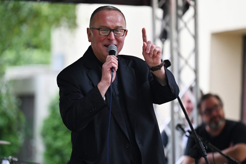 Ksiądz w czarnej marynarce i koloratce przemawia do mikrofonu na scenie, podnosząc palec w geście podkreślającym wypowiedź