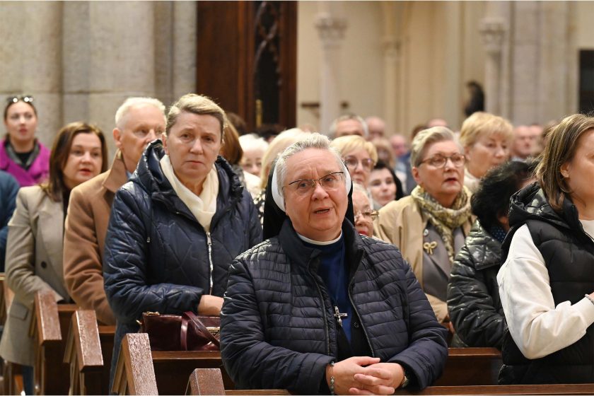 Fragment nawy z wiernymi stojącymi podczas modlitwy. Na pierwszym planie siostra zakonna w granatowym welonie i pikowanej kurtce, dłonie splecione; za nią kobiety w kurtkach i płaszczach, część w okularach, jedna w szaliku z roślinnym wzorem. Wszyscy zwróceni ku ołtarzowi, światło jasne, naturalne. Wrażenie jedności i skupienia.