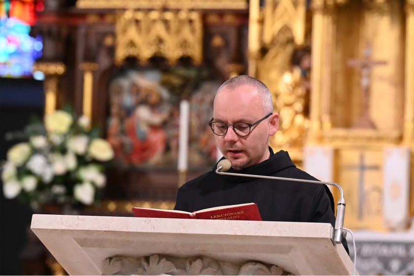 Zakonnik w czarnym habicie i okrągłych okularach czyta przy ambonie z jasnego kamienia. Przed nim otwarty czerwony „Lekcjonarz mszalny”, mikrofon na elastycznym ramieniu. W tle złocony ołtarz z detalami i rozmyte kwiaty. Kadr stabilny, podkreślający czynność czytania Słowa.