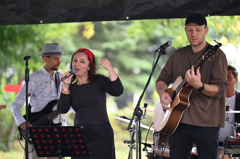 Zespół muzyczny na scenie – wokalistka z czerwoną opaską śpiewa do mikrofonu, obok gitarzysta gra na gitarze akustycznej, w tle perkusista i basista