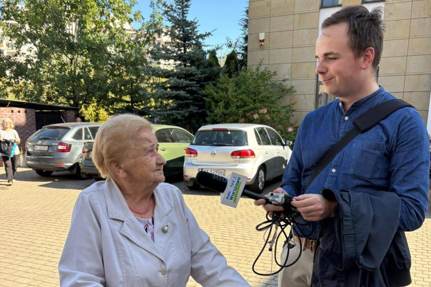 Dziennikarz z mikrofonem przeprowadza wywiad ze starszą kobietą przed budynkiem kościoła, w tle zaparkowane samochody i przechodnie