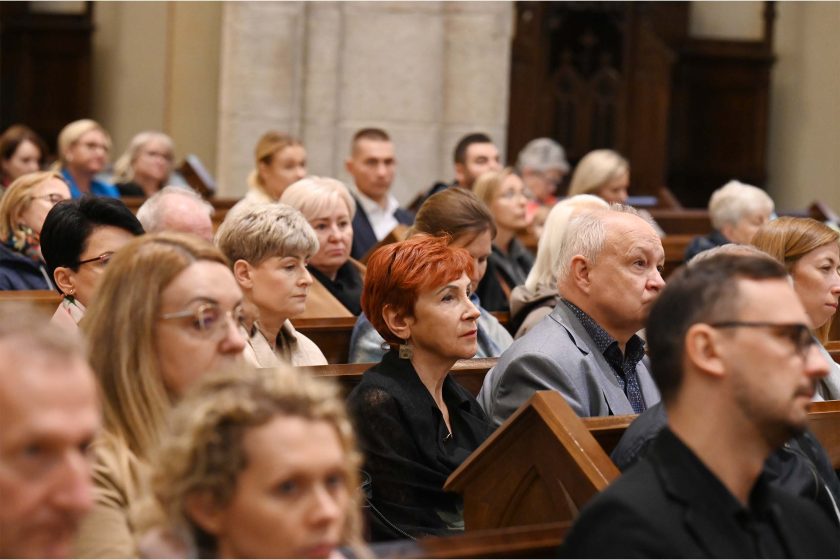 Wnętrze kościoła, ujęcie poziome na ławki z wiernymi. Na pierwszym planie, lekko z lewej, kobieta o krótkich rudych włosach i mężczyzna w szarej marynarce patrzą w stronę prezbiterium; za nimi osoby w różnym wieku, w płaszczach i okularach. Tło to kamienne filary i ciemne, drewniane elementy wystroju; światło miękkie, kościelne. Przód kadru delikatnie rozmyty, co kieruje uwagę na środkowy rząd ławek i skupione twarze.