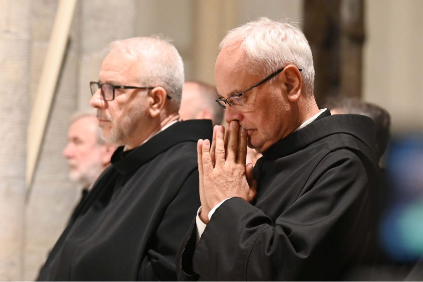 Półzbliżenie na dwóch zakonników w czarnych habitach, ujętych z profilu. Pierwszy, siwiejący, w okularach, patrzy przed siebie; drugi ma dłonie złożone i przyłożone do ust w geście cichej modlitwy, oczy zamknięte. Tło miękko rozmyte – kolumny i elementy wnętrza kościoła. Światło boczne rysuje delikatne cienie na twarzach, podkreślając skupienie.
