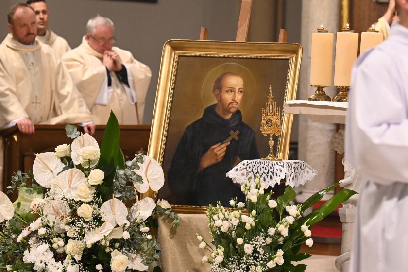 Ujęcie ołtarzowe: na sztalugach stoi obraz świętego zakonnika w złotej ramie; postać z nimbem trzyma mały krzyż w prawej dłoni. Obok, na koronkowej serwecie, złota, misternie zdobiona monstrancja-relikwiarz. Przed obrazem obfite białe kwiaty (m.in. anturia, róże, eukaliptus, gipsówka). W tle, po lewej, kilku duchownych w jasnych ornatach, nieostrych. Po prawej trzy kremowe świece na świeczniku. Atmosfera uroczysta, sakralna.